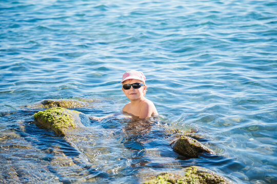 Blond boy aged five or six in striped cap and black sunglasses swims in clear blue sea near mossy rocksю