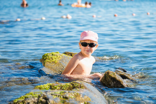 Boy aged 5 to 6 in pink striped cap and sunglasses sits between mossy rocks in clear sea water.