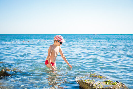Blond boy aged five or six in striped cap and black sunglasses swims in clear blue sea near mossy rocksю