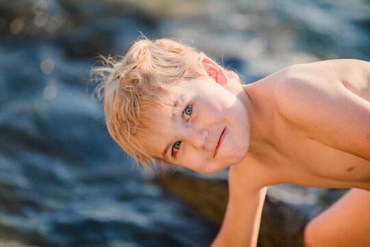 Close sunlit portrait of blond boy aged 5 to 6 with soft smile by sparkling sea rocks.
