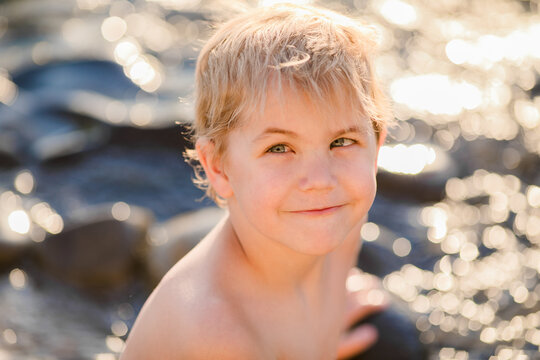 Close sunlit portrait of blond boy aged 5 to 6 with soft smile by sparkling sea rocks.