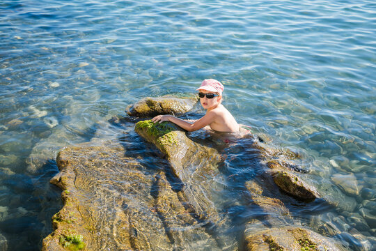 Blond boy aged 5 to 6 in striped cap and dark sunglasses looks down while standing among sea rocks.