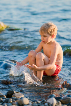 Boy aged 5 to 6 splashing water with his hands while sitting on seaside rocks.