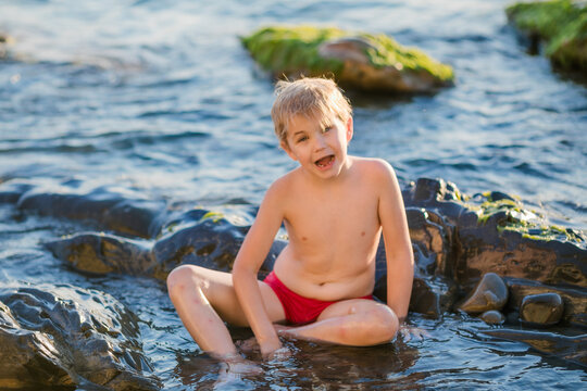 Smiling boy aged 5 to 6 sitting on wet sea rocks during a sunny summer day.