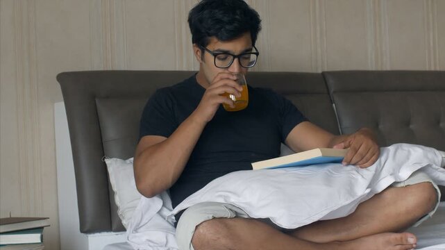 Young college student reading a book while sitting in his bed - lifestyle concept. Handsome Indian guy in glass spectacles reading a novel and having orange juice in his leisure time - hobby concept