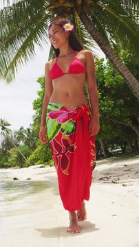 Tahiti luxury travel beach vacation woman walking in polynesian cover-up skirt beachwear on paradise island motu in French Polynesia. Red traditional clothes, bikini and flower. Video of real woman.