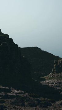 A rugged landscape shows tall rocky formations and uneven terrain. Low clouds cover the sky. The view captures a sense of vastness and natural elements.