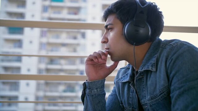 Young handsome man listening to music using his headphones - technology concept. Side view shot of a carefree Indian guy thinking something while sitting alone in his balcony - expressions concept