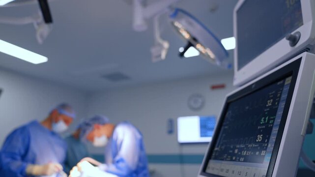 Footage at the monitor of lung ventilating machine working in surgery room. Three medics in blue uniforms perform operation in the blurred backdrop.