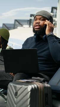 Vertical screen: African American manager takes a phone call in a casual outfit at the airport, handling work on the go beside runway windows during business travel.