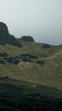 Groups of hikers move along a rocky path in a foggy area by the coast. The landscape features green hills and large stones. The atmosphere appears misty and cool.