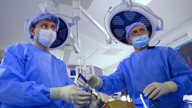Focused male doctors in blue uniforms stand near the patient holding a tool input into the incision. Low angle view. Modern round lamps in the backdrop.