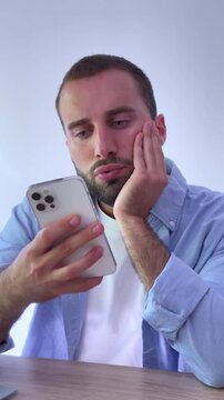 Sleepy young man with a beard looking bored while scrolling through social media feeds on his smartphone, feeling tired and uninterested while sitting at his desk at home
