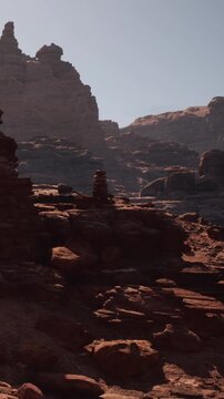 The scene shows tall rock formations in a desert environment. Sunlight shines on the rugged terrain. The background reveals more rocks and hills.