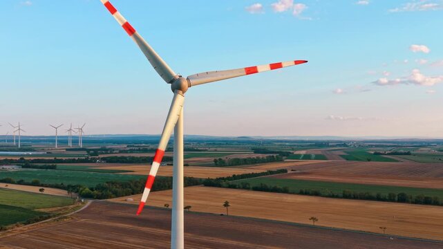 Aerial close view of wind turbine rotor and nacelle. A top down perspective highlights a modern generator over a colorful layout of harvested and green farmlands.