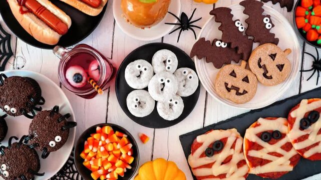Halloween party food table scene over a white wood background. Top view. Slow rotating zoom motion. Spooky mummy pizzas, finger hot dogs, cupcakes, candy, cookies and donuts.