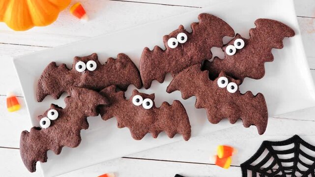 Plate of chocolate Halloween bat cookies. Top view with decor on a white wood background. Slow rotating zoom motion.
