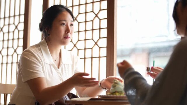 Woman smiles, conversing with friend at cafe. Hands gesture warmly, showing engagement. Window frames softly lit background. Calm, friendly interaction, perfect for lifestyle or social media