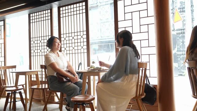 Two women converse at a wooden table. They sit near large windows with geometric latticework. One smiles, the other listens attentively. The scene suggests casual, friendly conversation