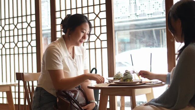 Two women share a cheerful moment at a cozy cafe. One smiles warmly while the other gestures animatedly. Soft light filters through patterned windows behind them. Their conversation feels relaxed