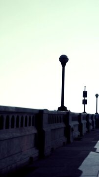 People stroll along a walkway beside the water. A streetlight stands nearby. A traffic sign is visible. The time is late evening. The sky has a soft light.