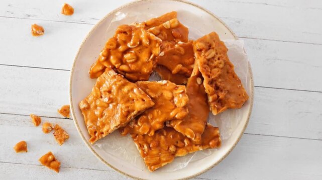 Plate of traditional peanut brittle candy pieces. Top view on a white wood background. Slow rotating zoom motion.