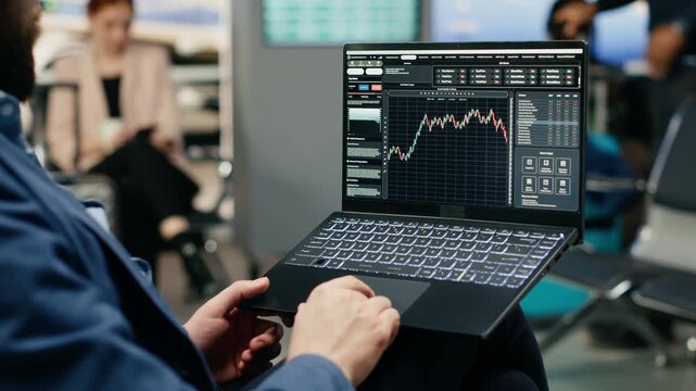 A business delegate monitors trading data and stock exchange graphs on a laptop in an airport lounge, tracking financial forecasts and investment trends while waiting near the gate.