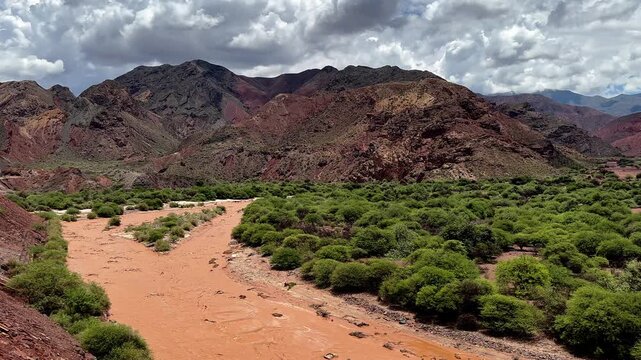 Panoramic view of a muddy river in a dry valley with green shrub and red mountains in Salta, Argentina. Scenic landscape, vivid, saturated colors. South America 