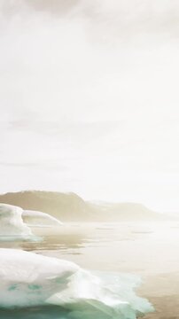 Icebergs are visible in the water while mountains are seen in the distance. The sun is setting, creating a warm glow over the scene. Calm waters reflect the surroundings clearly.