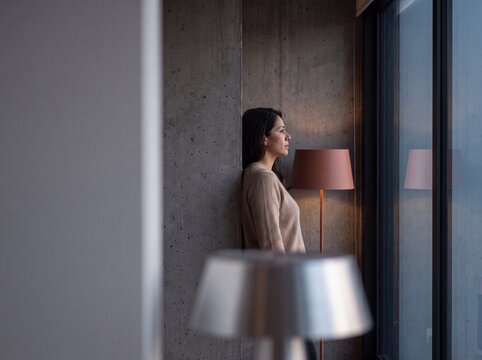 Pensive woman leaning against a raw concrete wall while thoughtfully looking out of a modern apartment window