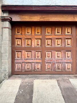 Geometric brown wooden double door with raised square panels and heavy lintel, surrounded by stone wall and tiled floor, traditional colonial architecture in Mexico.
