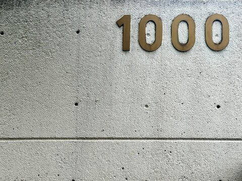 Number 1000 in golden metal digits on a grey architectural concrete wall. Minimalist background with house number on a rough cement surface.