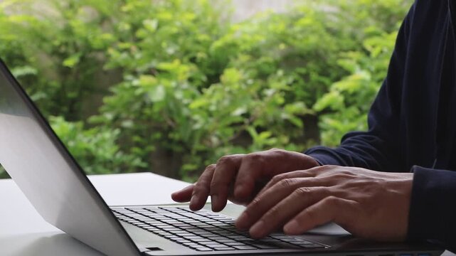 Close up male hands of unrecognizable male man at table typing computer keyboard online. Man typing on a laptop while sitting in the garden. Internet technologies concept.