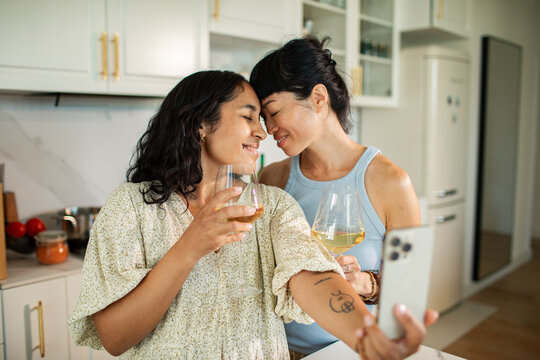 Same-sex couple taking wine selfie in home kitchen