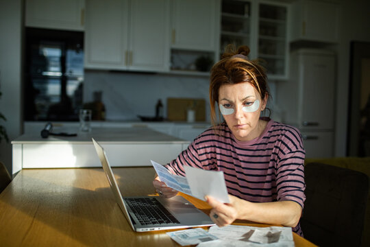 Tired woman reviewing bills with laptop at home kitchen table