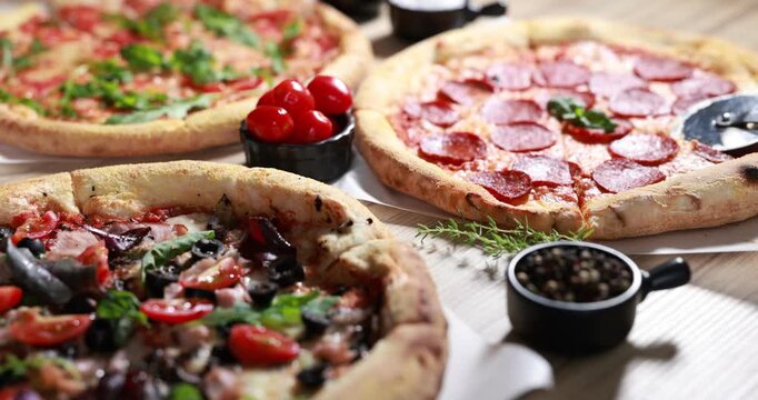 Different yummy pizzas, ingredients and cutter on wooden table, closeup. Camera moving