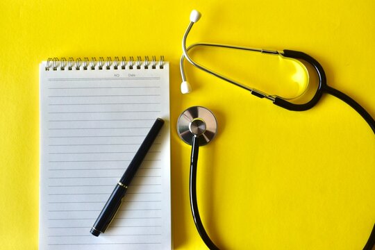 Top view of doctor workspace featuring a black pen, empty spiral notepad, and a diagnostic stethoscope on a yellow surface.