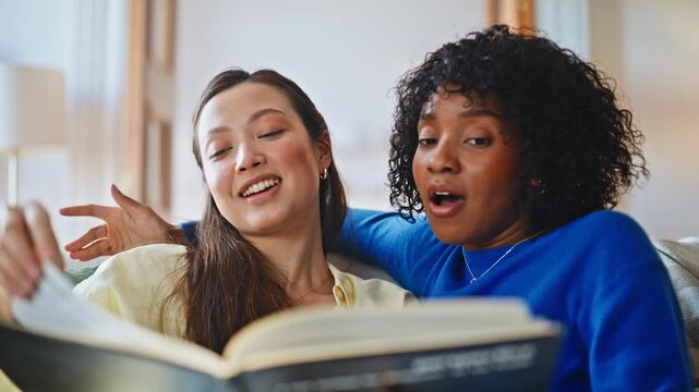 Two women reading book on sofa. Close up multiracial pair sharing story home.