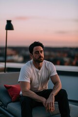 Fototapeta premium Pensive man in white t-shirt sitting on rooftop at sunset