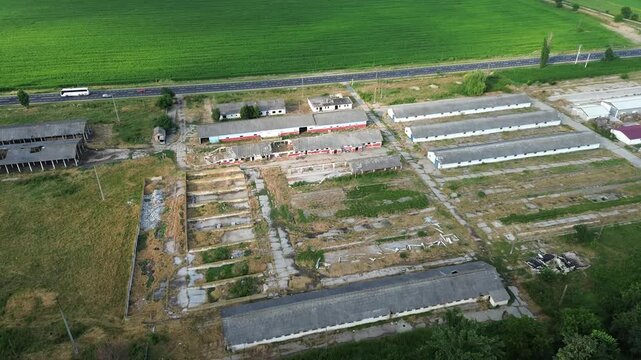 Aerial drone view of an abandoned agricultural farm complex featuring multiple long industrial buildings, deteriorated infrastructure, and open concrete yards surrounded by green farmland, nearby road