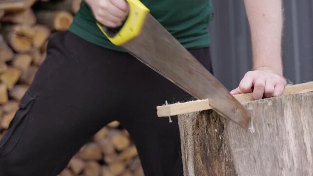 Unrecognizable young man in green shirt sawing piece of wood with manual hand saw on wooden block outdoors close up side view of motion and movement of carpenter at work in backyard on sunny day clip.