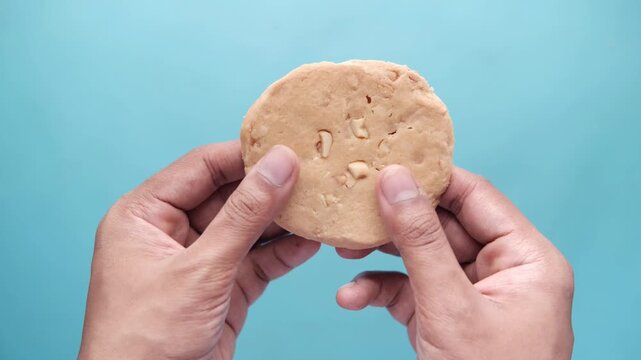 Close up of person breaking cashew cookie on light blue background, highlighting crispy texture and crumbs falling, perfect for food and snack related visual projects
