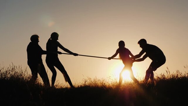 A happy family having fun playing tug of war at sunset, slow motion, a team of people pulling a rope in the park, sunlight through green grass.