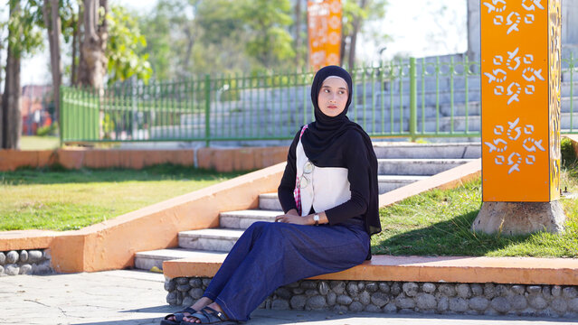 Young Asian Muslim Woman in Black Hijab Sitting on Outdoor Park Stairs Looking Away Thoughtfully in Modest Fashion Attire