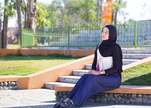 Young Asian Muslim Woman in Black Hijab Sitting on Outdoor Park Stairs Looking Away Thoughtfully in Modest Fashion Attire