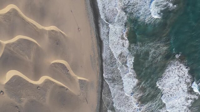An aerial drone shot of the stunning Maspalomas Dunes on Gran Canaria, featuring golden sand dunes meeting the turquoise ocean. The top down push in view shows the vast landscape and crashing waves.