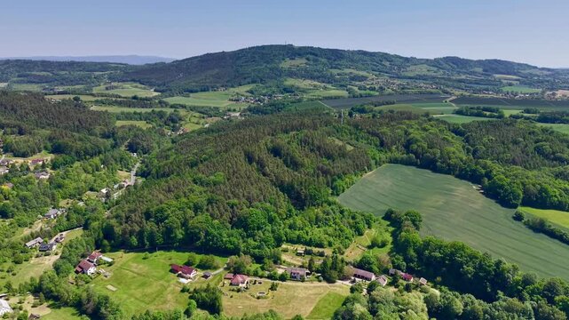 High oblique view moves over tranquil valley, wooded slopes, green fields, small houses, and open farmland, capturing soft shadows, wide scenery, calm rural landscape beneath clear blue sky