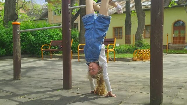 A medium shot captures a happy young girl in denim overalls smiling while hanging upside down from a metal playground bar during an inverted handstand trick, enjoying outdoor recreational play.