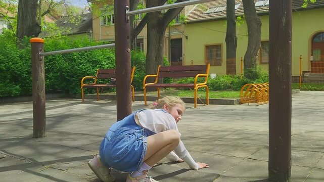 A medium shot captures a young girl in denim overalls performing an inverted handstand while hooked onto a metal pull-up bar at an outdoor park, preparing to dismount safely onto her feet.