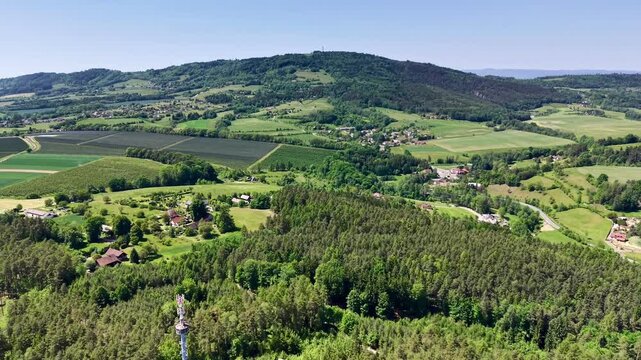 Smooth oblique aerial motion reveals broad rural valley with dense mixed forest in foreground, orchards, patchwork farmland, scattered farmhouses, small village, and wooded ridge under clear daylight